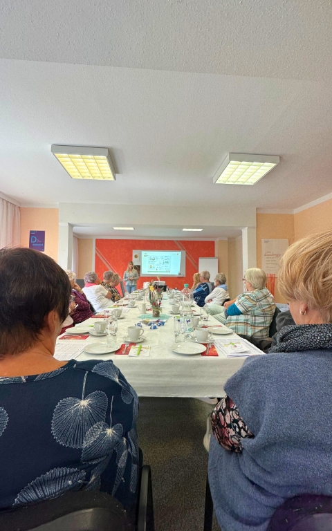 Das Foto zeigt eine große Tafel, an welcher 15 Personen sitzen. Alle Personen kehren sich mit dem Rücken zur Kamera und blicken Richtung Ende des Raumes. Dort steht eine Frau und hält einen Vortrag. 