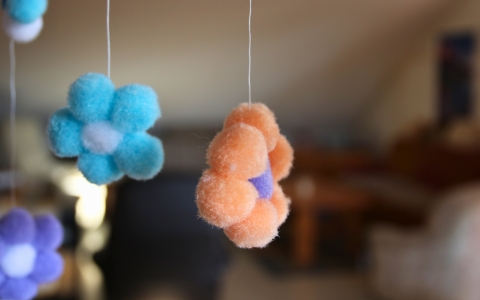 Im Vordergrund hängen mehrere flauschige, blumenförmige Objekte an dünnen Fäden. Die Blumen sind in verschiedenen Farben gestaltet, darunter Blau, Orange und Lila. Der Hintergrund ist unscharf und zeigt einen Innenraum mit Möbeln.