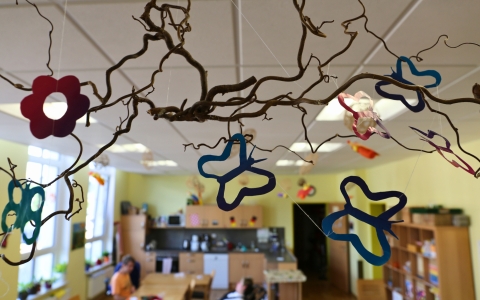 Ein Blick in einen freundlich eingerichteten Gruppenraum mit gelben Wänden, mehreren Fenstern und Deckenleuchten. Im Vordergrund hängen dekorative, bunte Papier- und Kunststoffformen in Blumen- und Schmetterlingsmotiven an einem geschwungenen Ast von der Decke, befestigt mit transparenten Fäden. Im Hintergrund sind Regale mit Spielsachen, eine Küchenzeile und ein großer Tisch mit mehreren Personen zu erkennen, die gemeinsam arbeiten oder basteln. Der Raum wirkt lebendig und kreativ, ideal für Gruppenaktivitäten und pädagogische Arbeit.
