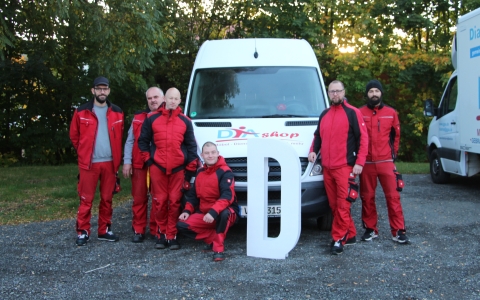 Sieben Personen in roten Overalls stehen und hocken vor einem weißen Transporter. Auf dem Transporter ist das Logo und der Schriftzug eines Unternehmens zu sehen. Eine große, weiße Buchstabenform steht vor dem Fahrzeug. Im Hintergrund sind grüne Bäume und ein weiterer Transporter zu erkennen.