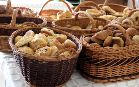 Mehrere geflochtene Brotkörbe aus braunem Weidengeflecht stehen auf einem mit einem karierten Tuch bedeckten Tisch. Die Körbe sind mit verschiedenen Brötchen und Backwaren gefüllt, darunter helle und dunkle Sorten sowie einige mit Körnern. Die Szene wirkt hell und einladend.