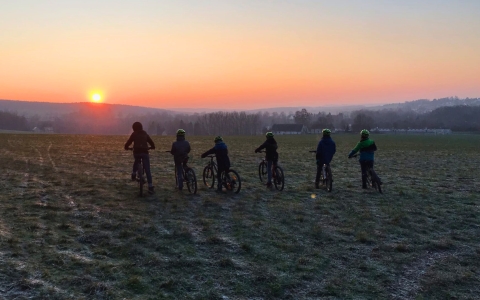 Das Bild zeigt sechs Kinder in Reihe nebeneinander auf Fahrrädern. Alle sind mit dem Rücken zur Kamera gedreht und blicken auf einen Sonnenuntergang. Alle Kinder tragen einen grünen Helm. Sie stehen auf einem Feld. Im Hintergrund ist eine hügelige Landschaft mit Sonnenuntergang. Es ist Dämmerung.