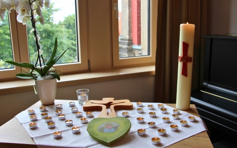 Auf einem kleinen Tisch vor einem Fenster stehen eine große Kerze mit rotem Kreuz, ein Holzkreuz, zahlreiche kleine Teelichter in Metallhüllen, ein grünes Herz mit einer gemalten Figur sowie eine weiße Orchidee in einem Blumentopf. Die Teelichter sind auf einem weißen Tuch angeordnet. Im Hintergrund sind Bäume und ein Gebäude durch das Fenster zu sehen. Rechts steht ein dunkles Klavier.