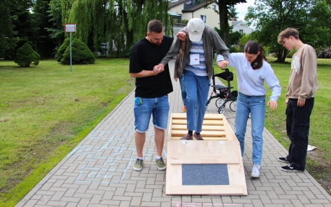 Das Foto zeigt vier Personen. Ein älterer Mann geht frontal über eine kleine Holzbrücke. Er schaut nach unten auf seine Füße. Er trägt um seinen Hals eine Art von Stempelkarte. Dieser Mann wird links von einem jungen Mann gestützt und rechts von einem Mädchen. Neben dem Mädchen steht noch ein Junge, welcher von der Seite aus zuschaut. Alle haben einen freudigen Gesichtsausdruck. Im Hintergrund sieht man einen gepflasterten Weg und einen Park. außerdem befindet sich hinter der Holzbrücke ein Rollator. Das wetter ist gut.