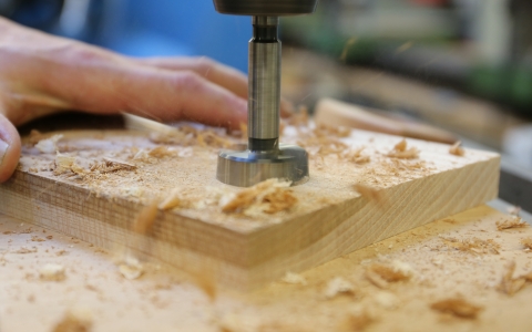 Eine Hand hält ein Holzbrett, auf das mit einer Standbohrmaschine ein großes Loch gebohrt wird. Rund um das Werkstück liegen Holzspäne, die beim Bohren entstanden sind. Im Hintergrund ist ein unscharfer Werkstattbereich zu sehen.