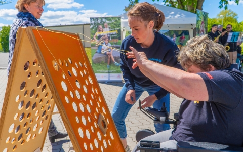 Im Vordergrund steht eine Person im Rollstuhl, die mit beiden Händen an einer gelben Schnur zieht. Die Schnur ist an einer großen, schräg stehenden Holzplatte befestigt, die zahlreiche runde Öffnungen aufweist. Zwei weitere Personen stehen daneben und beobachten die Szene. Im Hintergrund sind ein Plakat mit zwei Kindern, ein weißes Zelt und mehrere Menschen zu sehen. Die Umgebung ist sonnig, der Himmel ist blau mit wenigen Wolken.
