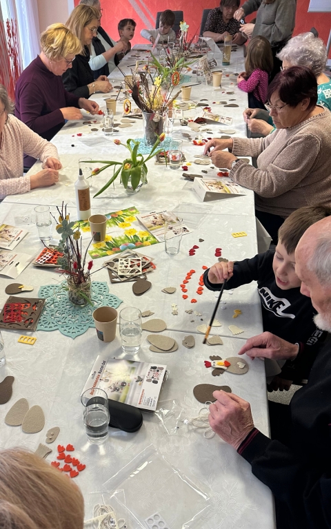 Auf dem Bild sieht man eine lange Tafel, an welcher unterschiedliche Menschen sitzen, sowohl Kinder als auch ältere Menschen. Alle sitzen zusammen und basteln. Auf dem Tisch stehen Blumen und es liegt viel Material zum Basteln durcheinander. 