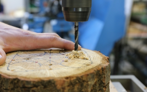 Eine Hand fixiert ein quer liegendes Stück Baumstamm, während mit einer Standbohrmaschine ein Loch in das Holz gebohrt wird. Auf der Oberfläche sind Markierungen und Linien zu sehen, die vermutlich Bohrpunkte oder Schnittführungen anzeigen. Im Hintergrund ist eine Werkstattatmosphäre mit Werkzeugen und Maschinen sichtbar.