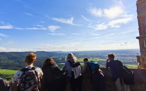 Das Foto zeigt fünf Jugendliche von hinten. Alle tragen Rücksäcke. Sie stehen an einer Mauer und blicken auf eine Landschaft. Die Landschaft ist weitläufig mit Wäldern und Siedlungen. Der Himmel ist sonnig.