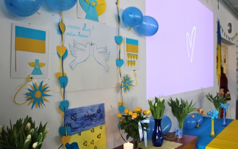 Wand mit blau-gelben Papierherzen, Luftballons, Zeichnungen von Tauben und ukrainischer Flagge. Auf einem Tisch stehen Blumenvasen mit Tulpen.