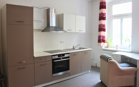 Küchenzeile mit hellbraunen und weißen Fronten, integriertem Backofen, Cerankochfeld und Dunstabzugshaube aus Edelstahl. Links ein hoher Schrank, rechts eine Spüle mit Unterschrank. An der Wand ein Fenster mit rot-weiß gestreiftem Vorhang, davor ein Schreibtisch mit einem Korbsessel. Auf dem Fensterbrett stehen mehrere grüne Pflanzen.