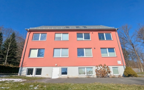 Das Foto zeigt ein großes rotes Haus mit acht Fenstern. Es ist frontal und von unten fotografiert. Im Hintergrund ist blauer Himmel und Wald. Im Vordergrund ist Wiese. 