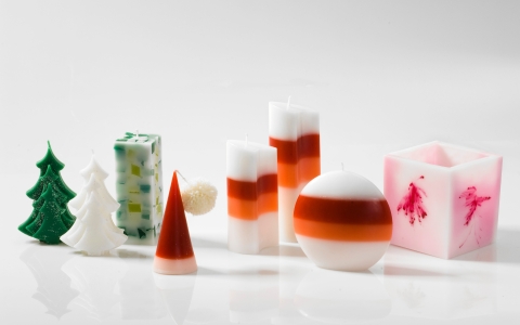 Verschiedene Kerzen stehen nebeneinander auf einer glatten, weißen Oberfläche. Die Kerzen haben unterschiedliche Formen und Farben: Eine grüne Kerze in Form eines Tannenbaums, eine weiße Kerze in Form eines Weihnachtsbaums, eine rot-weiße Kerze in Form einer Spitze mit einer kleinen weißen Kugel, zwei zylindrische Kerzen mit roten und weißen Streifen, eine kugelförmige Kerze mit roten und weißen Streifen, eine rechteckige Kerze mit grünen und blauen Einschlüssen sowie eine würfelförmige Kerze mit rosa und roten Mustern.