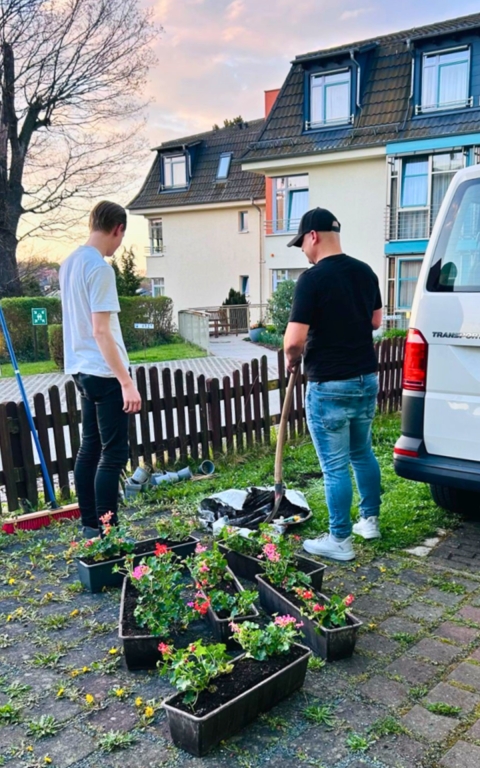 Zwei junge Männer arbeiten im Vordergrund gemeinsam an der Gestaltung eines kleinen Gartens: Einer hält einen Spaten, der andere schaut ihm zu. Beide stehen auf einem gepflasterten Bereich, der von mehreren Blumenkästen mit bunten Blütenpflanzen umgeben ist. Rechts im Bild steht ein weißer Van, dahinter sind Wohnhäuser mit gepflegten Vorgärten und Bäumen zu sehen. Ein niedriger Holzzaun trennt das Gartengrundstück von den Häusern. Die Szene findet zur Abendstunde statt, der Himmel ist hell mit warmen Farbtönen, was eine ruhige und gemütliche Stimmung vermittelt.
