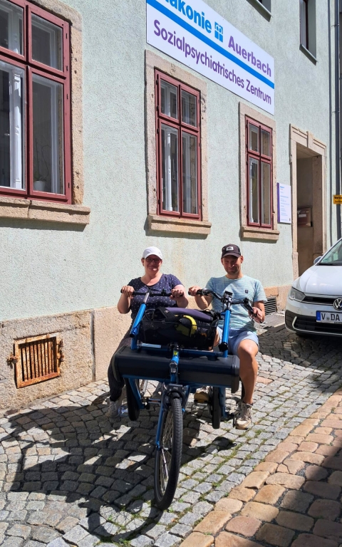 Zwei Personen sitzen nebeneinander auf einem blauen Spezialfahrrad mit drei Rädern. Beide tragen Freizeitkleidung und haben die Hände an den Griffen. Das Fahrrad steht auf einem Kopfsteinpflaster vor einem hellen Gebäude mit roten Fensterrahmen. Über ihnen hängt ein Schild mit der Aufschrift 'Diakonie Auerbach Sozialpsychiatrisches Zentrum'. Rechts im Bild steht ein weißer Pkw am Straßenrand.