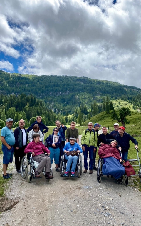 Das Bild zeigt eine Gruppe an Menschen, welche in die Kamera lächeln. Drei Personen sitzen im Rollstuhl. Alle tragen Outdoor- bkleidung oder Wanderkleidung. Im Hintergrund ist eine bergige und grüne Landschaft zu sehen mit Wald. Im Vordergund befindet sich ein sandiger Weg. Das Wetter ist wolkig und sonnig.
