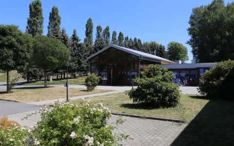 Außenaufnahme eines modernen, eingeschossigen Gebäudes mit großen Fenstern und einem schrägen Dach inmitten einer gepflegten Grünanlage. Im Vordergrund befinden sich blühende Büsche und ein gepflasterter Weg, der zur Eingangstür des Gebäudes führt. Die Umgebung ist von Bäumen umrahmt und vermittelt einen freundlichen, offenen und naturnahen Eindruck bei strahlendem Sonnenschein und blauem Himmel.