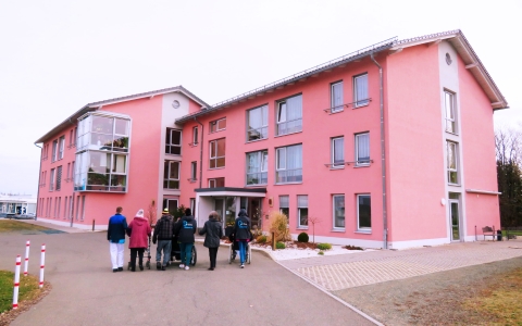 Im Hintergrund des Bildes sieht man ein großes rotes Haus, mit vielen Fenstern. Das Haus sieht einladend aus. Im Vordergrund sieht man eine Reihe aus Menschen, welche auf das Haus zugehen. Man sieht den Rücken der Personen. Einige von Ihnen tragen eine Di