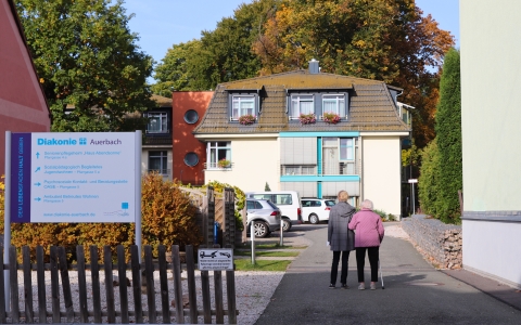 Auf dem Bild ist ein gelbes Haus zu sehen. Im Hintergrund ist ein herbstlicher Park. Im Vordergrund sieht man zwei Frauen auf das Haus zulaufen. Die eine Frau stützt die andere. Im Vordergrund links ist eine Infotafel zu sehen. Auf dieser sind verschieden