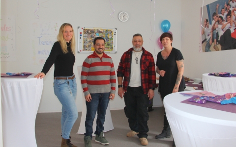 Drei Frauen und ein Mann blicken in die Kamera, sie stehen in einem Raum mit Stehtischen, Luftballons und Wanddekorationen