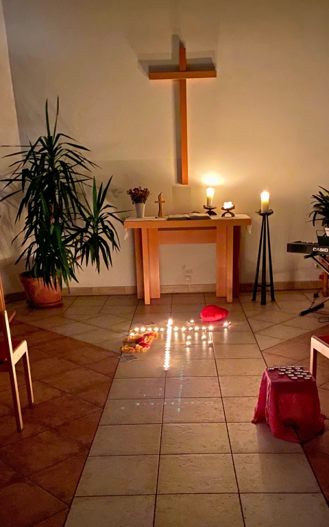 Ein schlichter Altarraum mit einem Holzkreuz an der Wand, flankiert von zwei großen Grünpflanzen in Töpfen. Auf dem Altar stehen Kerzen, eine Bibel und ein Blumengefäß. Vor dem Altar sind zahlreiche Teelichter in einer Linie auf dem Boden angeordnet, daneben liegen ein rotes Stoffherz und einige bunte Blätter. Rechts im Bildrand ist ein Teil eines Keyboards zu sehen, links und vorne stehen einfache Holzstühle. Die Szene ist warm beleuchtet und vermittelt eine ruhige, besinnliche Atmosphäre.