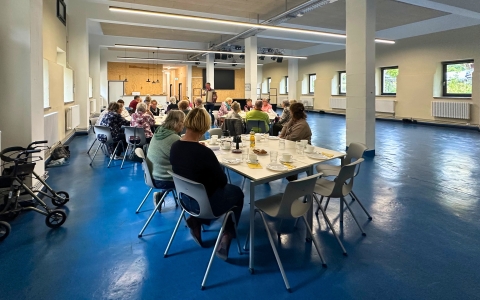 Das Bild zeigt einen großen Saal mit mehreren Tischen. An diesen sitzen viele Personen. Auf den Tischen stehen Kaffeegedecke. Im Hintergrund des Saals sieht man einen Mann, welcher Akkordeon spielt. Alle Personen schauen fröhlich zu dem Musiker.