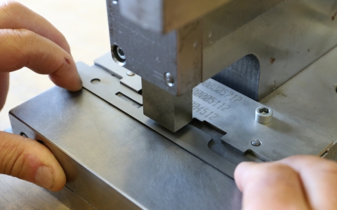 Eine Metallplatte wird mit einer Maschine bearbeitet. Zwei Hände halten die Platte fest, während ein massiver Metallstempel von oben auf das Werkstück drückt. Die Oberfläche der Maschine und der Platte ist glatt und silbern. Auf der Metallplatte sind Zahlen und Buchstaben eingraviert.