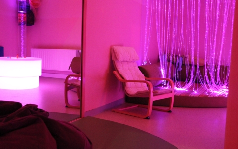Ein gemütlicher, in rosa und lila Licht getauchter Raum, ausgestattet mit einem weißen Sessel und einer weichen Matte. Im Hintergrund hängen viele leuchtende Lichtfasern, die für eine entspannende Atmosphäre sorgen, während eine beleuchtete Blasensäule einen weiteren Sinnesanreiz bietet. Der Raum wirkt ruhig und lädt zum Wohlfühlen ein.