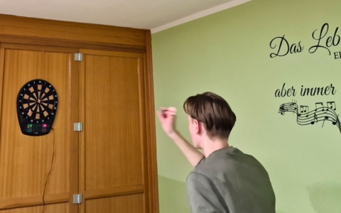 Ein junger Mensch mit hellbraunen Haaren steht vor einer hellgrünen Wand und wirft einen Dartpfeil auf eine elektronische Dartscheibe, die an einer Holzschranktür befestigt ist. An der Wand befindet sich ein dekorativer Schriftzug mit Musiknoten.
