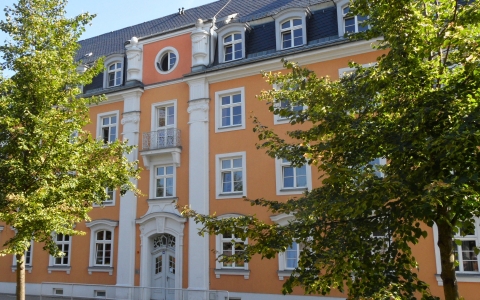 Das Bild zeigt ein großes gelb- orangenes Gebäude mit weißen Fenstern- und Fensterrahmen.   Mittig ist eine große weiße Tür zu sehen und darüber ein weißer Eisen-Balkon. Das Haus sieht sehr einladend aus. Das Haus wird links und rechts im Vordergrund von grünen Bäumen verdeckt. Der Himmel ist blau. 