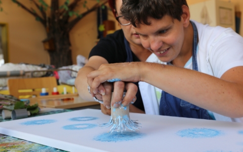 In einem Kreativraum sitzen zwei Personen an einem Tisch. Eine Person wird dabei von einer anderen angeleitet und unterstützt: Zusammen führen sie eine Hand, die mit blauer Farbe bestrichen ist, auf eine weiße Leinwand und stempeln mit den Fingern kreisförmige Muster. Im Hintergrund sind bunte Malutensilien wie Farben und Pinsel sowie Dekorationen und Möbelstücke zu erkennen. Die Szene vermittelt kreatives, gemeinschaftliches Arbeiten und künstlerische Förderung.