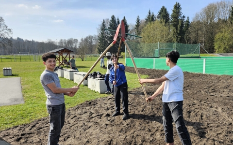 Drei Personen stehen auf einem umgegrabenen Erdstreifen im Freien. Jede Person hält eine lange Schaufel, die Stiele treffen sich in der Mitte und tragen ein rotes Spatenblatt. Im Hintergrund sind eine grüne Umzäunung, ein Fußballplatz, Bäume und eine Sitzecke zu sehen. Die Personen tragen Freizeitkleidung.