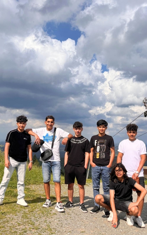 Eine Gruppe von sechs Jugendlichen posiert im Freien auf einem Hügel oder Aussichtspunkt. Sie stehen und hocken locker nebeneinander, schauen in die Kamera und wirken entspannt. Im Hintergrund sind eine weite Landschaft und ein dramatischer Himmel mit großen, dichten Wolken zu sehen.