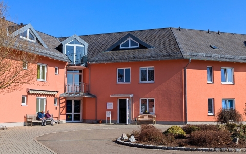 Das Foto zeigt ein rotes Haus. In der Mitte befindet sich eine große Glastür. Neben dieser rechts ist eine weiter Tür, welche offen steht. Davor ist auch ein Briefkasten und eine Bank. Im Bild links sitzen auch zwei Herren auf einer Bank vor dem Haus in der Sonne. Im Vordergrund des Hauses befindet sich ein großes rundes Beet. Der Himmel im Hintergrund ist blau.