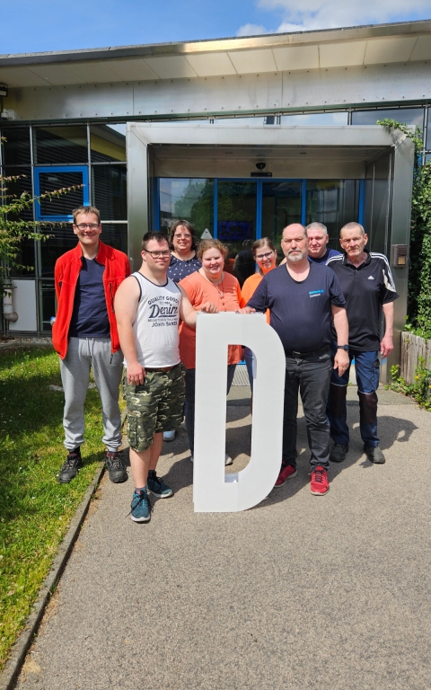 Eine Gruppe von Menschen steht im Freien vor einem Gebäude mit großen Glasfenstern und blauen Türen. Die Personen halten gemeinsam ein großes, weißes, freistehendes D aus festem Material. Die Kleidung ist unterschiedlich, darunter T-Shirts, Shorts, Jeans und Sportbekleidung. Der Hintergrund zeigt einen gepflasterten Weg, etwas Grün und einen Teil des Gebäudes.