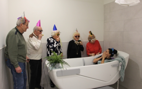 Auf dem Bild ist eine Badewanne zu sehen. In dieser sitzt eine Frau mit erschrockenem Gesichtsausdruck und hält sich die Hände vor den Mund. Um die Badewanne herum stehen drei Frauen und zwei Männer, welche auf die Badewanne schauen. Sie tragen alle einen
