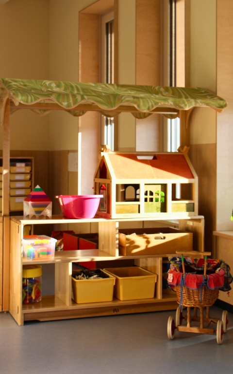 Im Vordergrund steht ein Holzregal mit mehreren offenen Fächern, in denen gelbe und rote Kunststoffkisten sowie Spielzeug verstaut sind. Auf dem Regal befindet sich ein großes Puppenhaus aus Holz mit grünen Fenstern und einer gelben Tür. Darüber ist ein Baldachin mit grünem Stoffdach angebracht. Rechts neben dem Regal steht ein Puppenwagen aus geflochtenem Material mit einem rot karierten Stoffbezug. Im Hintergrund sind hohe Fenster und beige gestrichene Wände zu sehen. Das Sonnenlicht fällt durch die Fenster und beleuchtet den Raum.