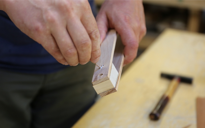 Eine Person hält ein Holzstück in der einen Hand, während sie mit der anderen Hand einen Nagel in das Holz führt. Das Holzstück hat eine eckige Form, und auf dem Arbeitstisch liegt ein Hammer. Im Hintergrund ist eine Werkstattumgebung leicht unscharf erkennbar.