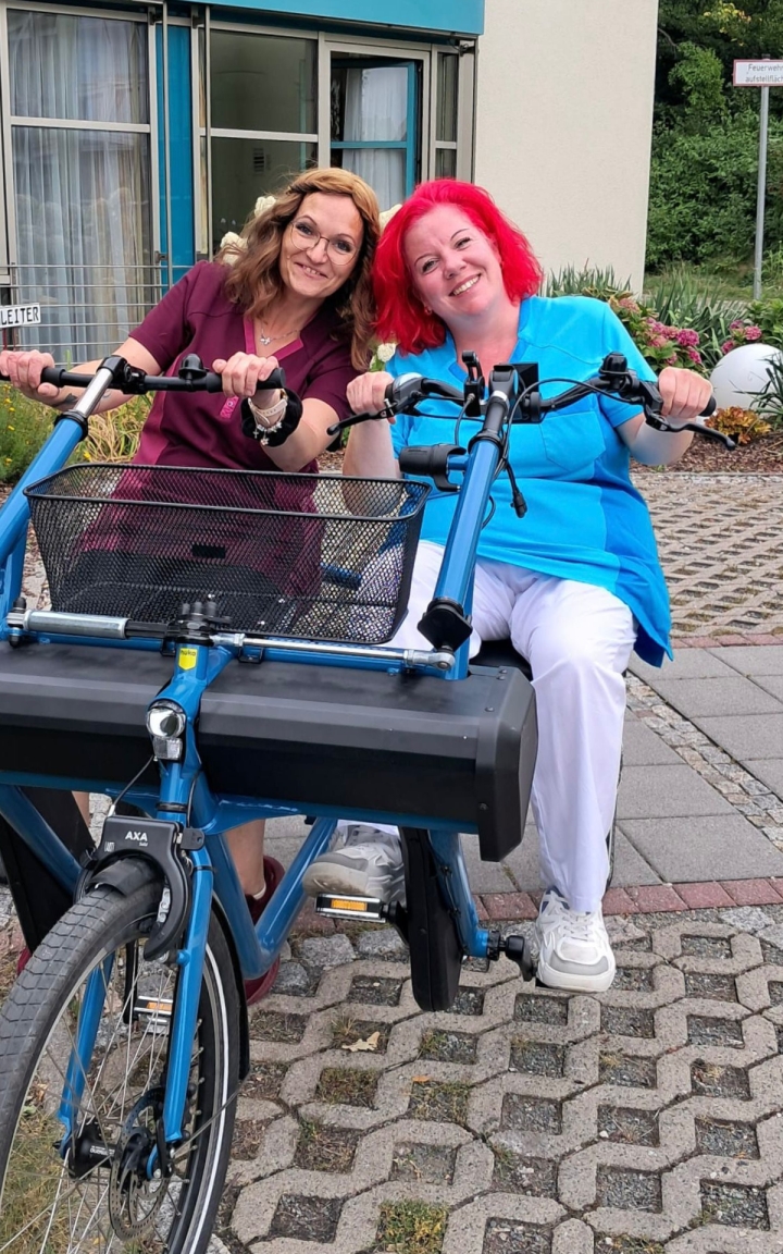 Auf dem Bild sind zwei Mitarbeiterinnen zu sehen, welche auf einem speziellen Fahrrad nebeneinander sitzen. Beide Frauen lächeln in die Kamera.  si