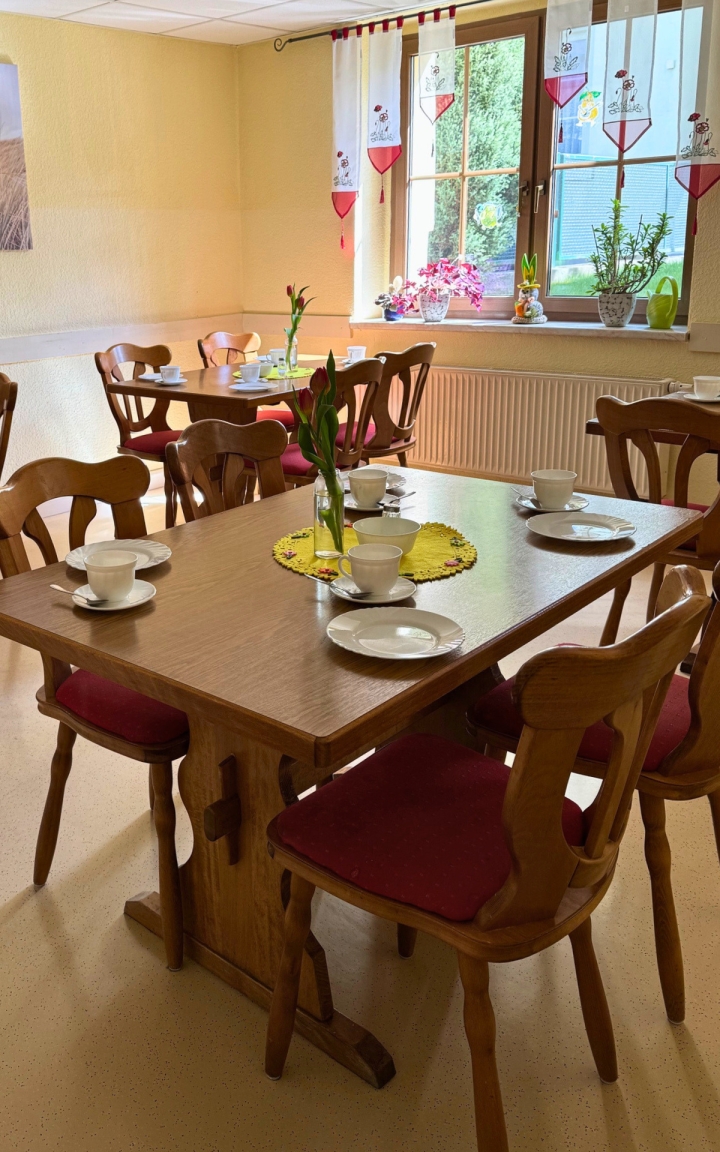 Zu sehen ist ein gemütlicher, kleiner Speiseraum mit mehreren rechteckigen Holztischen und Stühlen mit rot gepolsterten Sitzflächen. Auf jedem Tisch stehen Gedecke mit Tassen, Untertassen und kleinen Tellern, arrangiert für eine gemeinsame Mahlzeit; vereinzelt befinden sich kleine Blumenväschen und dekorative Mitteldecken. Die Fensterwand im Hintergrund lässt Tageslicht ins Zimmer, auf der Fensterbank befinden sich weitere Blumen und Dekorationen. Die raumhohen Vorhänge sind weiß mit roten Herzen und freundlichen Motiven. Das Gesamtbild vermittelt eine einladende, wohnliche Atmosphäre.