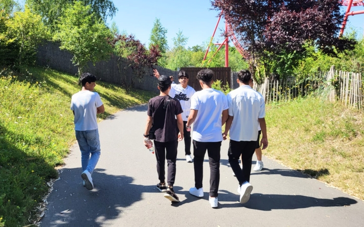 Eine Gruppe von sechs jungen Männern läuft an einem sonnigen Tag gemeinsam einen asphaltierten Weg entlang, umgeben von viel Grün und Bäumen. Im Hintergrund ist eine rote Achterbahnstruktur sichtbar. Einer der Männer dreht sich zur Gruppe um und scheint etwas zu sagen oder zu zeigen, während die anderen ihm folgen.