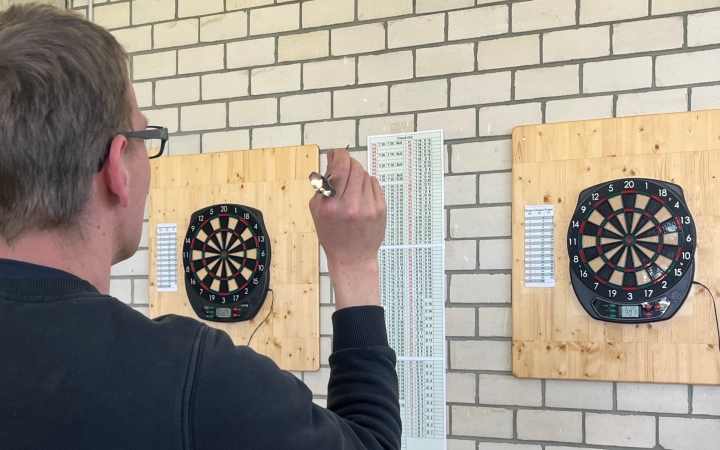 Ein Mann steht an einer Wand mit hellen Ziegeln, auf der zwei elektronische Dartscheiben auf Holzbrettern montiert sind. Er hält einen Dartpfeil in der rechten Hand und zielt auf die linke Scheibe. Daneben hängen Spielpläne oder Tabellen. Im Bildvordergrund ist der Mann von hinten zu sehen, er trägt eine Brille und dunkle Kleidung. Die Szene zeigt den Moment vor dem Wurf beim Darts.