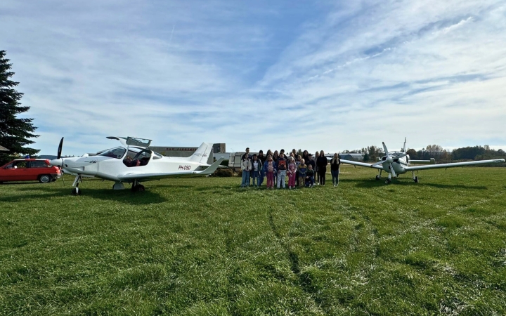 Zwei kleine, weiße Sportflugzeuge stehen auf einer Wiese. Dazwischen steht eine Gruppe von Menschen in bunter Freizeitkleidung. Im Hintergrund sind ein rotes Auto, ein Gebäude und Bäume zu sehen. Der Himmel ist klar und blau.