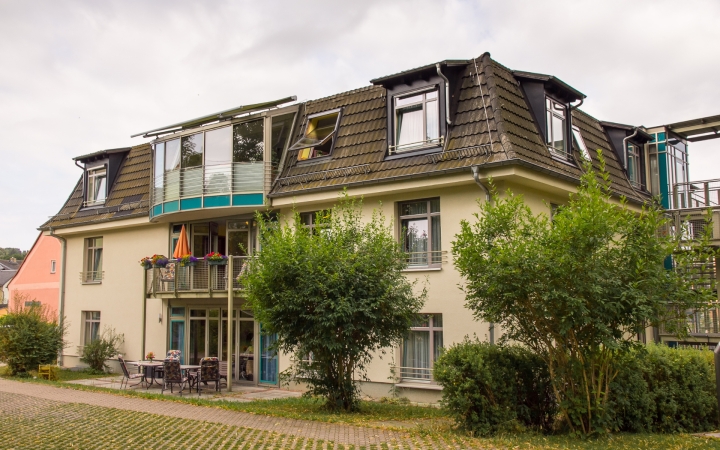 Auf dem Bild ist ein gelbes Haus zu sehen. Das Haus hat viele Fenster und einen Balkon und eine Terrasse. Im Vordergrund rechts wird das Haus von grünen Büschen verdeckt.