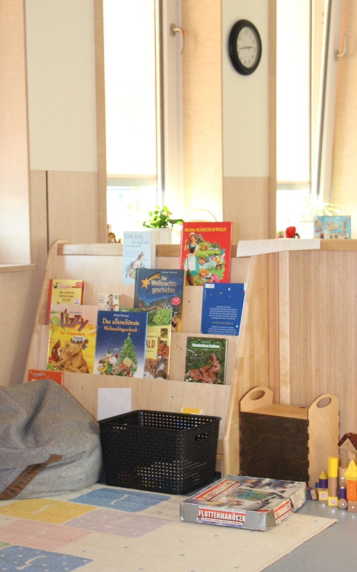 Eine gemütliche Leseecke in einem hellen Raum mit großen Fenstern. Ein Bücherregal aus Holz steht im Mittelpunkt, gefüllt mit verschiedensten Kinderbüchern, deren Buchcover sichtbar sind. Neben dem Regal steht ein grauer Sitzsack, eine schwarze Kiste und ein Brettspiel („Flottenmanöver“) auf einem bunten Teppich mit geometrischen Mustern. In der Ecke sind Holzspielzeuge und weitere Elemente zu sehen. Die Uhr an der Wand zeigt ungefähr 10 Uhr. Das Licht fällt sanft durch die Fenster und es stehen einige Pflanzen auf dem Fensterbrett.