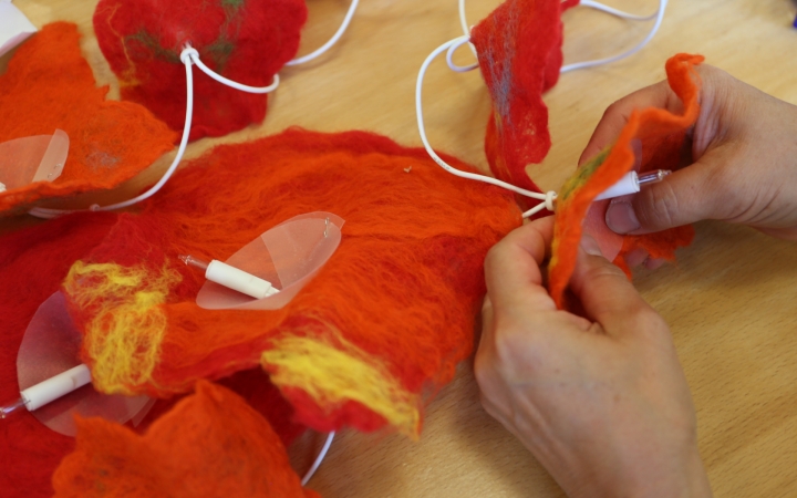 Auf einem Holzarbeitstisch werden mehrere dekorative, leuchtend rote und orangefarbene, handgefertigte Filzformen arrangiert und an weiße Kabel montiert. Eine Person ist zu sehen, wie sie die Filzelemente über einen Lampensockel stülpt und befestigt. Die Szene vermittelt das Basteln oder Montieren einer kreativen Lichterkette in filigraner Handarbeit. Die Filzflächen wirken lebendig farbig und sind teilweise mit gelben Fasern durchsetz
