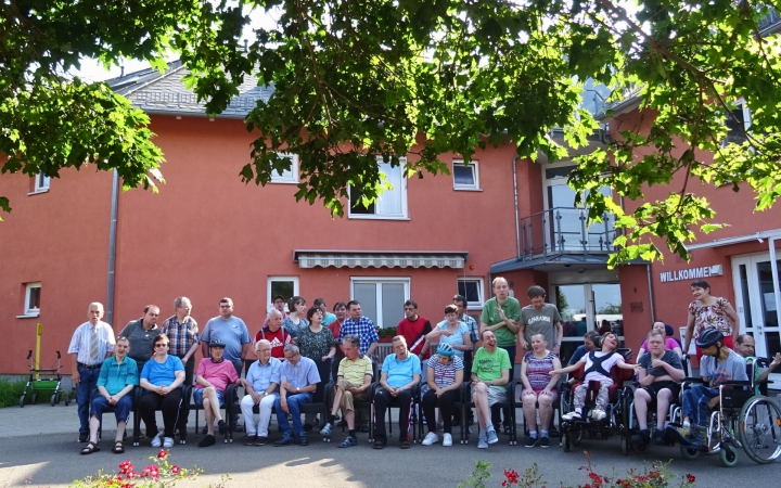 Das Foto zeigt circa 30 Menschen, welche alle neben- und hintereinander stehen und in die Kamera schauen. In der vorderen Reihe sitzen alle Personen auf einem Stuhl oder im Rollstuhl. Die weiteren Personen verteilen sich dahinter in die Lücken. Im Hintergrund sieht man ein rotes Haus, welches rechts im Bild die Aufschrift 