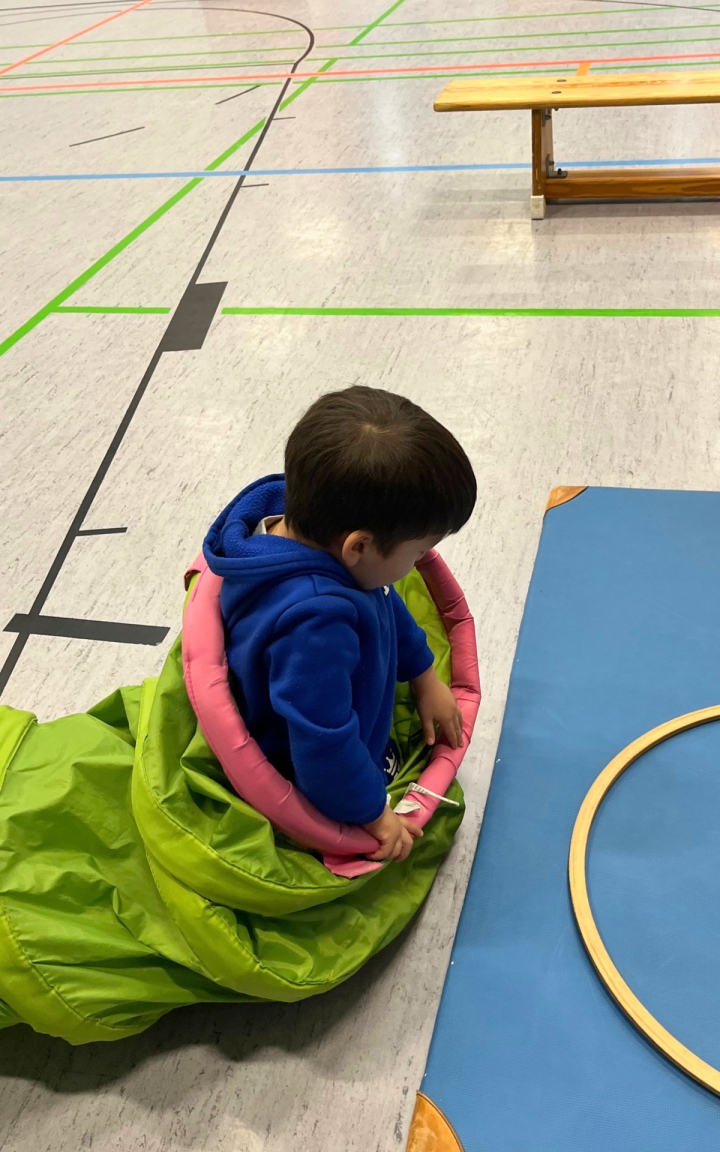 Ein Kind mit braunen Haaren sitzt auf dem Boden einer Turnhalle. Es trägt einen blauen Kapuzenpullover und ist teilweise in einen grünen Schlafsack oder eine Decke gehüllt. Um den Oberkörper liegt ein pinkfarbener Gymnastikreifen. Im Hintergrund sind eine blaue Matte, ein Holzreifen und eine Turnbank zu sehen. Der Boden ist mit bunten Linien markiert.