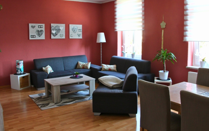 Wohnzimmer mit roten Wänden, zwei dunkelgrauen Sofas und einem hellen Holztisch auf einem gemusterten Teppich. Im Hintergrund stehen ein weißer Stehlampe und eine grüne Topfpflanze auf einem weißen Übertopf. An der Wand hängen vier gerahmte Bilder mit Herzmotiven. Zwei große Fenster mit halbtransparenten, gestreiften Rollos lassen Tageslicht herein. Im Vordergrund steht ein Esstisch mit vier gepolsterten Stühlen.