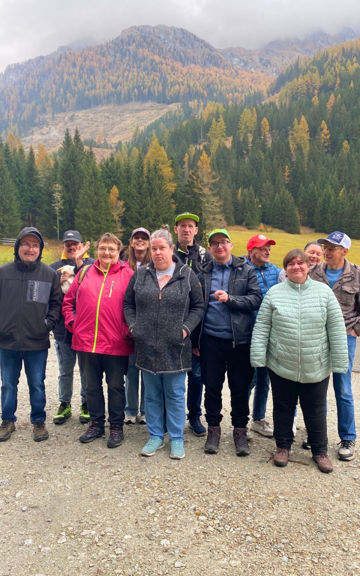 Das Foto zeigt eine Gruppe an unterschiedlichen Menschen. Alle lächeln zur Kamera. Die Personen targen alle Kleidung zum Wandern. Im Hintergrund ist eine herbstliche Landschaft zu sehen mit Bergen und Wäldern. Das Wetter ist wolkig und trüb.